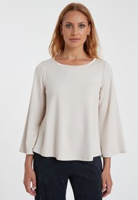 Lichtbeige langemouwen top met een ronde hals en uitlopende mouwen. Voordelige textuur, minimalistisch ontwerp en subtiele glans bij de halslijn.