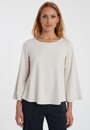 Helle beigefarbene Langarmbluse mit rundem Ausschnitt und ausgestellten Ärmeln. Glatte Textur, minimalistisches Design und dezent glänzender Ausschnitt.