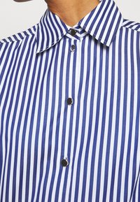 Camicia a righe blu e bianche, con colletto a punta e bottoni neri. Tessuto morbido, caratterizzato da righe verticali regolarmente distanziate.