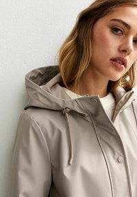 Veste imperméable beige avec capuche, dotée d'une fermeture éclair et d'accents à cordon. Texture douce et design minimaliste.