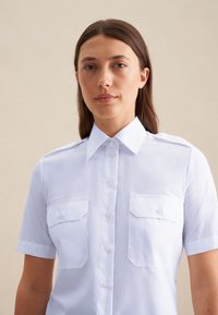 Camicia bianca a maniche corte con colletto a punta, due tasche sul petto, chiusura con bottoni e una texture liscia. Presenta maniche dalla linea dritta.