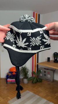 Gorro malhado a preto e branco com um desenho geométrico, pompom no topo e tranças pendendo dos lados.