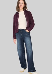 Chaqueta de botón borroso burdeos sobre una blusa crema clara, combinada con pantalones vaqueros de pierna ancha azul oscuro y zapatillas blancas con suelas beige.