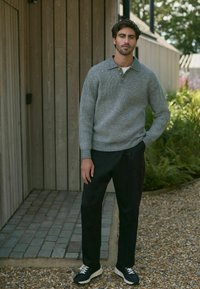 Suéter gris de punto con cuello, combinado con pantalones negros. El conjunto presenta unas zapatillas negras y blancas sobre un camino exterior texturizado.