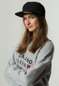 Căciulă snapback neagră cu vizieră plată, purtată cu un hanorac gri, care are un text îndrăzneț negru. Părul este lung și ondulat, iar brațele sunt încrucișate.