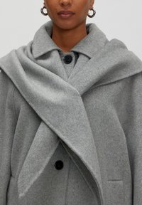 Grauer Wollmantel mit großem Kragen, mit Knopfverschluss und überlappendem Stoff, der vorne drapiert ist. Weiche Textur und minimalistisches Design.