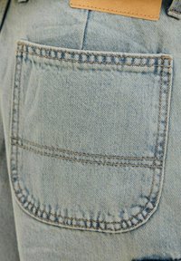 Poche de jean en denim bleu clair avec une double couture visible et un patch en cuir marron au-dessus du bord supérieur de la poche.