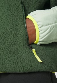 Zielona polarowa kurtka o teksturowanej powierzchni i gładkim, jasnozielonym rękawie. Posiada żółte akcenty oraz kieszeń na zamek.