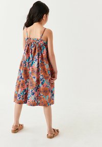 Jeune fille se tenant de dos, portant une robe sans manches à motif floral avec des couleurs orange, bleue et rose, ainsi que des sandales beiges.