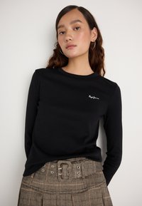 Fekete, hosszú ujjú póló bordázott textúrával és egy kis fehér logóval a mellrészen, amelyet egy fém részletekkel ellátott kockás szoknyával párosítanak.