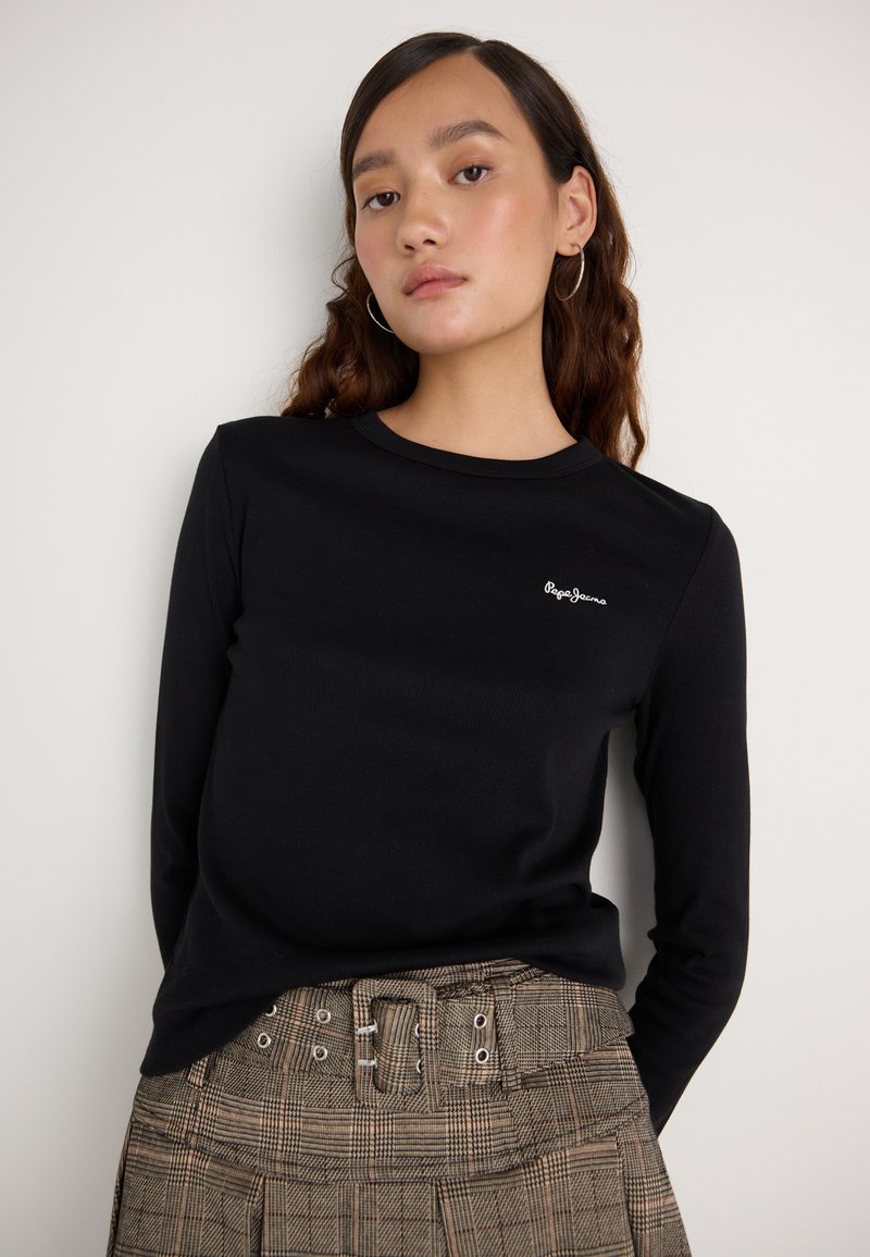 Fekete, hosszú ujjú póló bordázott textúrával és egy kis fehér logóval a mellrészen, amelyet egy fém részletekkel ellátott kockás szoknyával párosítanak.