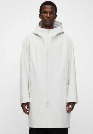 Mężczyzna w długim, białym, z kapturem płaszczu z przednim zamkiem błyskawicznym i bocznymi kieszeniami, stojący na jasnym, jednolitym tle.