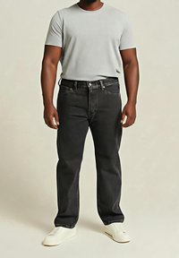 Homme portant un t-shirt à manches courtes gris clair, un jean noir et des chaussures blanches sans lacets, debout sur un fond uni.