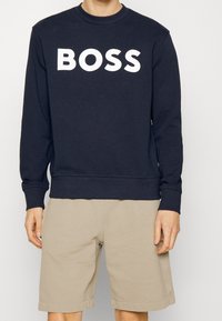 Námořnický modrý svetr z měkké látky s dlouhými rukávy a kulatým výstřihem, s velkým bílým logem "BOSS" přes prsa. V kombinaci s béžovými kraťasy.