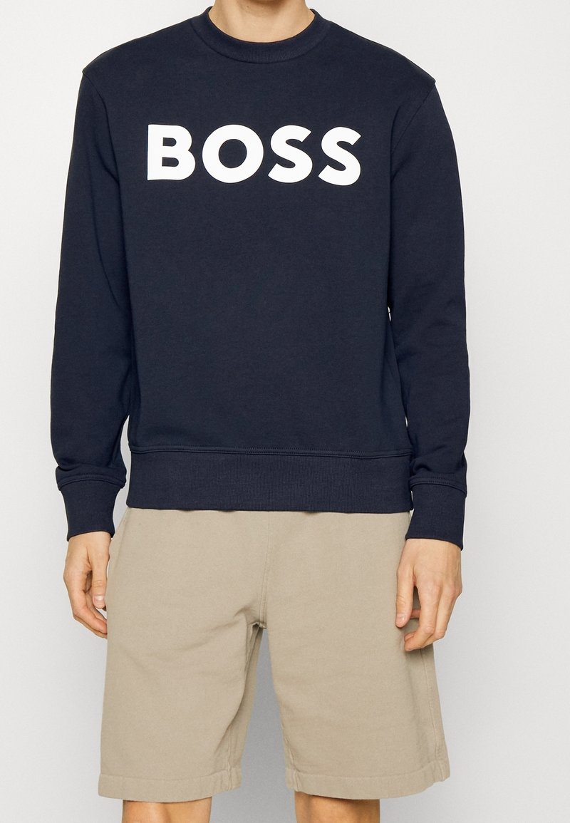 Námořnický modrý svetr z měkké látky s dlouhými rukávy a kulatým výstřihem, s velkým bílým logem "BOSS" přes prsa. V kombinaci s béžovými kraťasy.