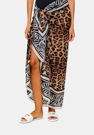 Donna che indossa un sarong con stampa leopardata e motivi ai bordi in bianco e nero legato in vita, abbinato a pantofole bianche aperte sulle punte con dettaglio a cuore dorato.