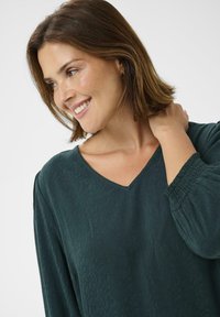 Femme aux cheveux bruns mi-longs portant un blouse vert foncé texturé avec des poignets smockés, souriant et touchant son cou, sur un fond blanc.