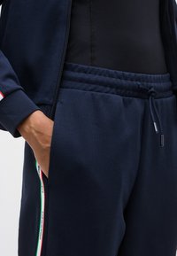 Άτομο που φοράει σκούρο μπλε παντελόνι φόρμας με κορδόνι στη μέση και πλαϊνές ρίγες σε πράσινο, λευκό και κόκκινο, με το χέρι μέσα στην τσέπη.