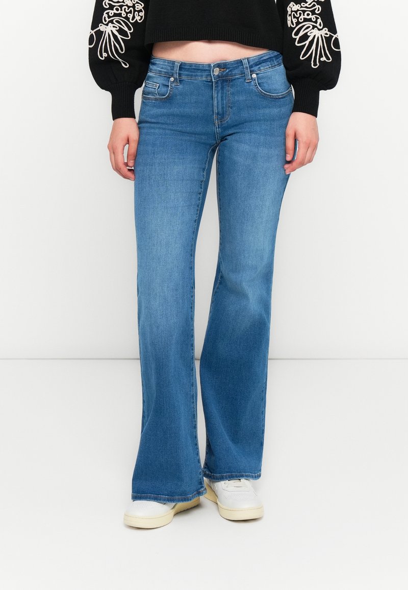 Jeans acampanados azules con una textura suave, lavado medio, diseño de cinco bolsillos y un leve desgaste a lo largo de la pierna, combinados con zapatillas blancas.