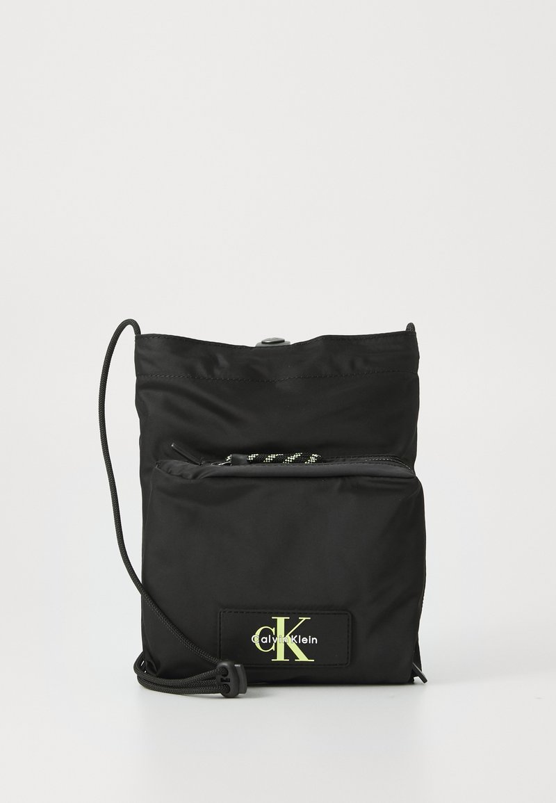 Svart nylon crossbody-veske med justerbar snorrem, frontlomme med glidelås, og fluorescerende logo-lapp på lærdetalj.
