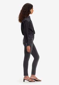 Camisa de mezclilla negra con cierre de botones y bolsillo en el pecho, combinada con jeans gris ajustados con acentos de tachuelas. Llevada con sandalias de tacón negras.