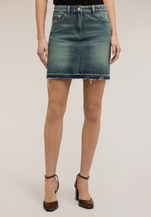 Kobieta w wyblakłej niebieskiej dżinsowej mini spódniczce z postrzępionym dołem i brązowych szpilkach na kostkę, stojąca na jednolitym tle.