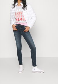 Kvinna som bär en vit vadderad jacka, grå och rosa grafisk "LOVE" sweatshirt, mörka smala jeans och vita höga Converse-sneakers.