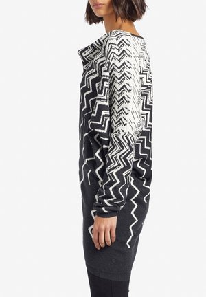 Donna che indossa un vestito lungo a maglione con motivo zigzag bianco e nero, con maniche lunghe, in piedi di profilo su uno sfondo bianco.