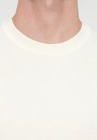 Bawełniany t-shirt w kolorze kremowym, z prążkowanym dekoltem w serek i gładką, miękką fakturą. Brak wzorów lub nadruków.