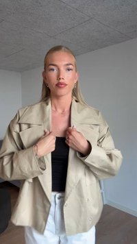 Beige übergroße Jacke mit großen Revers, Knopfdetails und glatter Textur, über einem schwarzen Top getragen, kombiniert mit weißen Hosen.