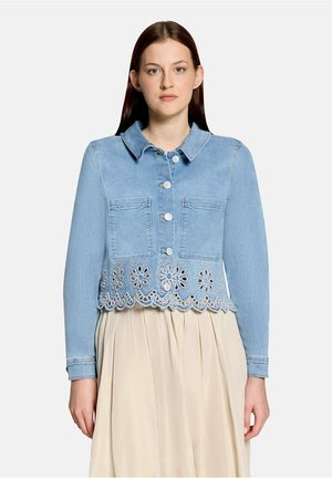 Vrouw draagt een lichtblauwe spijkerjasje met bloemvormige oogjes aan de zoom en witte knopen, gecombineerd met een beige geplooide rok.