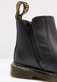 Black leather ankle boot with side zipper, textured finish, yellow stitching along the sole, and a pull tab with brand logo on top.
