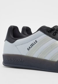 Az Adidas Gazelle tornacipők világosszürke textil felsőrésszel, fekete bőr részletekkel, három fehér csíkkal és érdes fekete gumitalppal rendelkeznek.