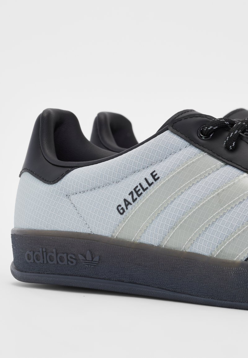 Die Adidas Gazelle Sneakers verfügen über ein hellgraues Textil-Obermaterial, schwarze Lederelemente, drei weiße Streifen und eine strukturierte schwarze Gummisohle.