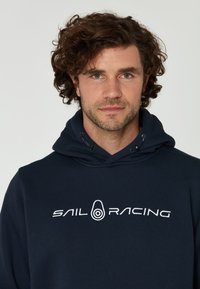 Námořnická modrá mikina s kapucí vyrobená z měkké tkaniny, se bílým logem "SAIL RACING" na hrudi, s nastavovací šnúrkou na kapuci a pohodlným střihem.