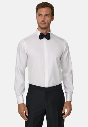 Un homme pose avec une chemise blanche habillée, un nœud papillon noir et un pantalon noir sur un fond clair et uni.