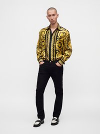 Homme debout, les mains dans les poches, portant une chemise à motifs dorés et noirs, un pantalon noir et des mocassins noirs et blancs, sur fond uni.