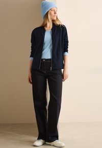 Giacca zip-up blu navale sopra una camicia azzurro chiaro, abbinata a jeans scuri e scarpe bianche. Un berretto azzurro chiaro con texture sulla testa aggiunge un tocco di stile.