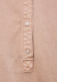 Tessuto beige con una texture liscia, caratterizzato da due bottoni decorativi argentati e dettagli cuciti lungo il fondo.