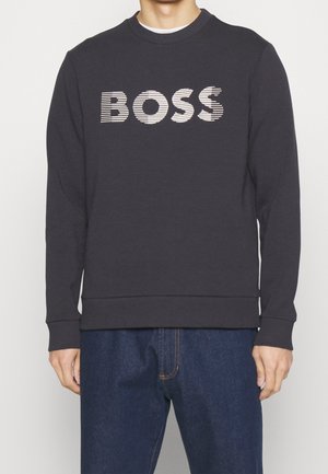 Mężczyzna ubrany w czarną, długorękawową bluzę z napisem "BOSS" w białe paski na piersi oraz w niebieskie dżinsy.
