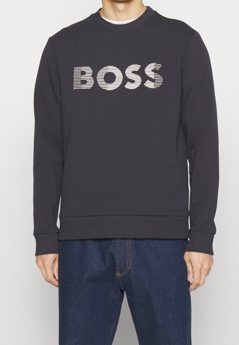 Homme portant un sweat-shirt noir à manches longues avec le texte rayé blanc "BOSS" sur la poitrine et un jean bleu.