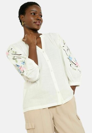 Mujer con blusa blanca abotonada con bordados florales coloridos en las mangas y pantalones cargo beige, mirando hacia su izquierda con la mano cerca del cuello.