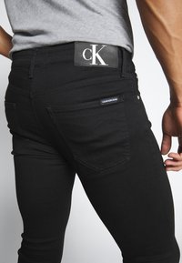 Acercamiento de una persona con jeans negros de Calvin Klein y una camisa gris, mostrando el bolsillo trasero y la etiqueta del logo en la cintura.