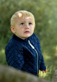 Niño con chaqueta acolchada azul oscuro con cremallera, mirando hacia arriba. Tiene el pelo rubio corto y parece estar al aire libre con vegetación de fondo.