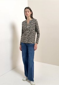 Chemise à manches longues avec un motif noir et beige et un col en V, associée à un jean à jambes larges bleu et des baskets blanches. Tenue simple et décontractée.
