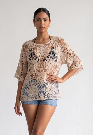 Femme portant un haut en crochet beige par-dessus un bikini noir et un short en jean bleu, debout avec une main sur la hanche devant un fond uni.