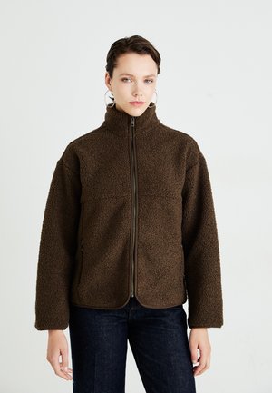 Femme portant une veste en polaire zipée marron foncé avec un col haut, un jean bleu et de grandes boucles d'oreilles contre un fond clair.
