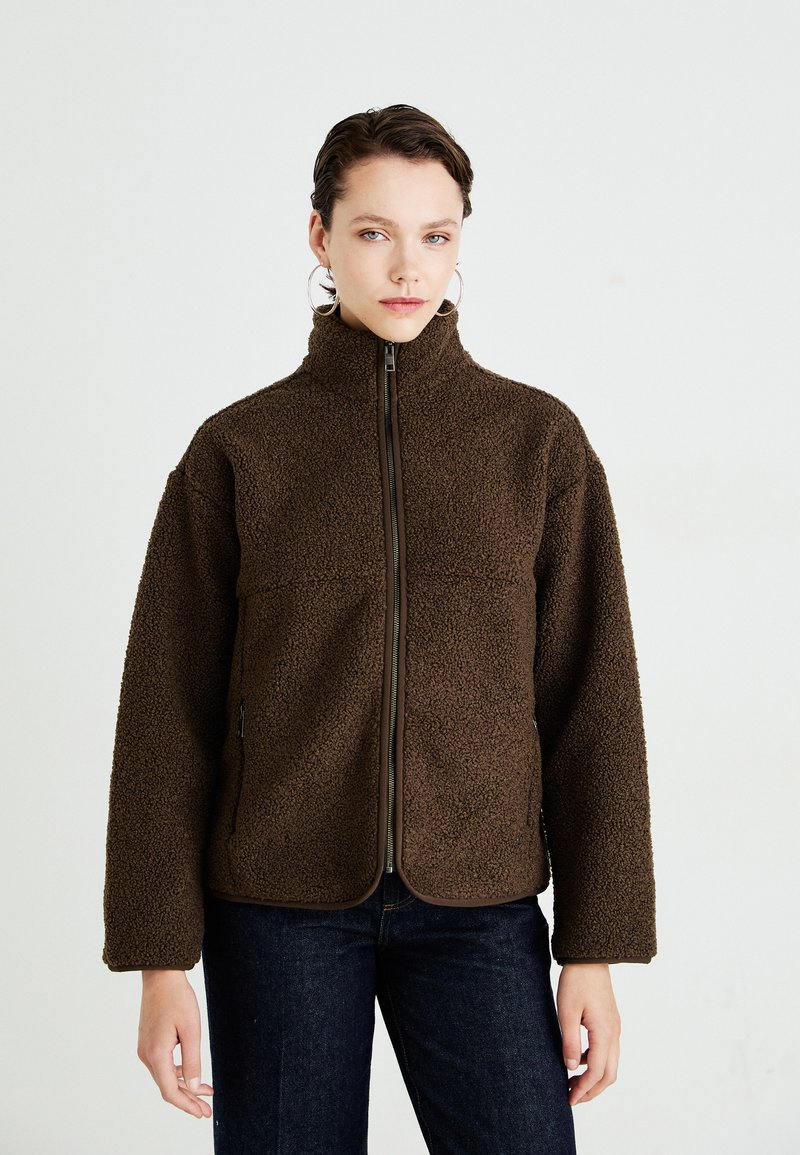 Woman wearing a dark brown zip-up fleece jacket with high collar, blue jeans, and large hoop earrings against a plain light background.