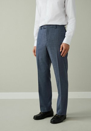 Muž nosiaci modré na mieru šité nohavice, bielu košeľu s dlhým rukávom a čierne kožené mokasíny, stojaci pred jednoduchým svetlým pozadím.