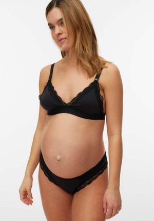 Femme enceinte portant un soutien-gorge de maternité noir bordé de dentelle et un sous-vêtement assorti, debout sur fond blanc.